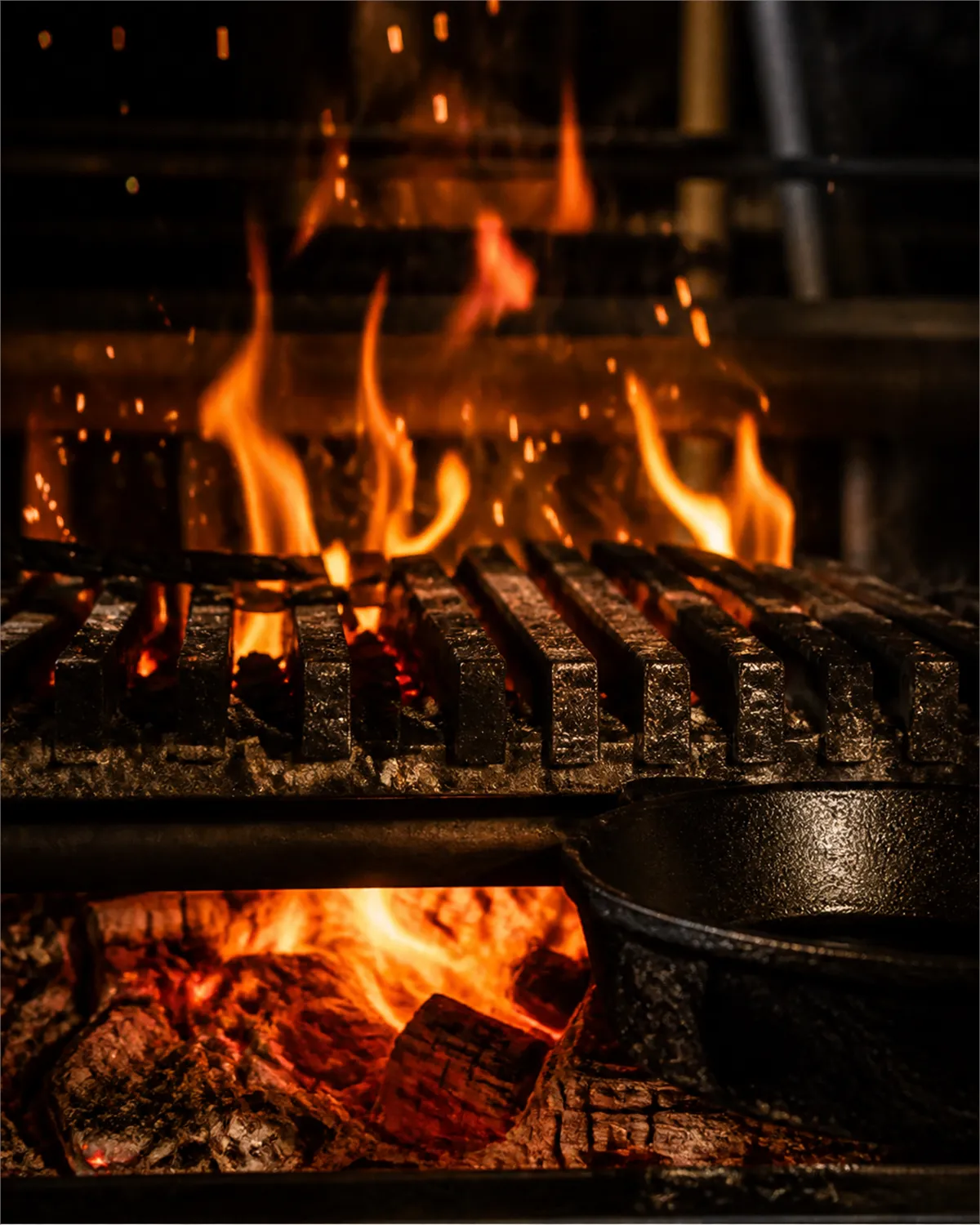 Flames rising through the bars of the oak grill, embers and split logs glowing below, a cast iron pan at the edge
