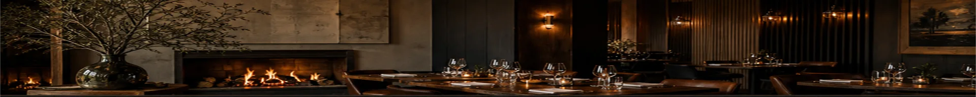 The dining room at Ember & Oak: linen-set tables, wine glasses catching the light, and the long open fireplace glowing along the wall